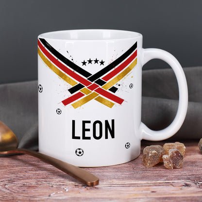 Personalisierte Fußball Tasse mit Name Leon, Fan-Design in Schwarz Rot Gold, kleine Fußbälle und Zuckerstücke im Hintergrund