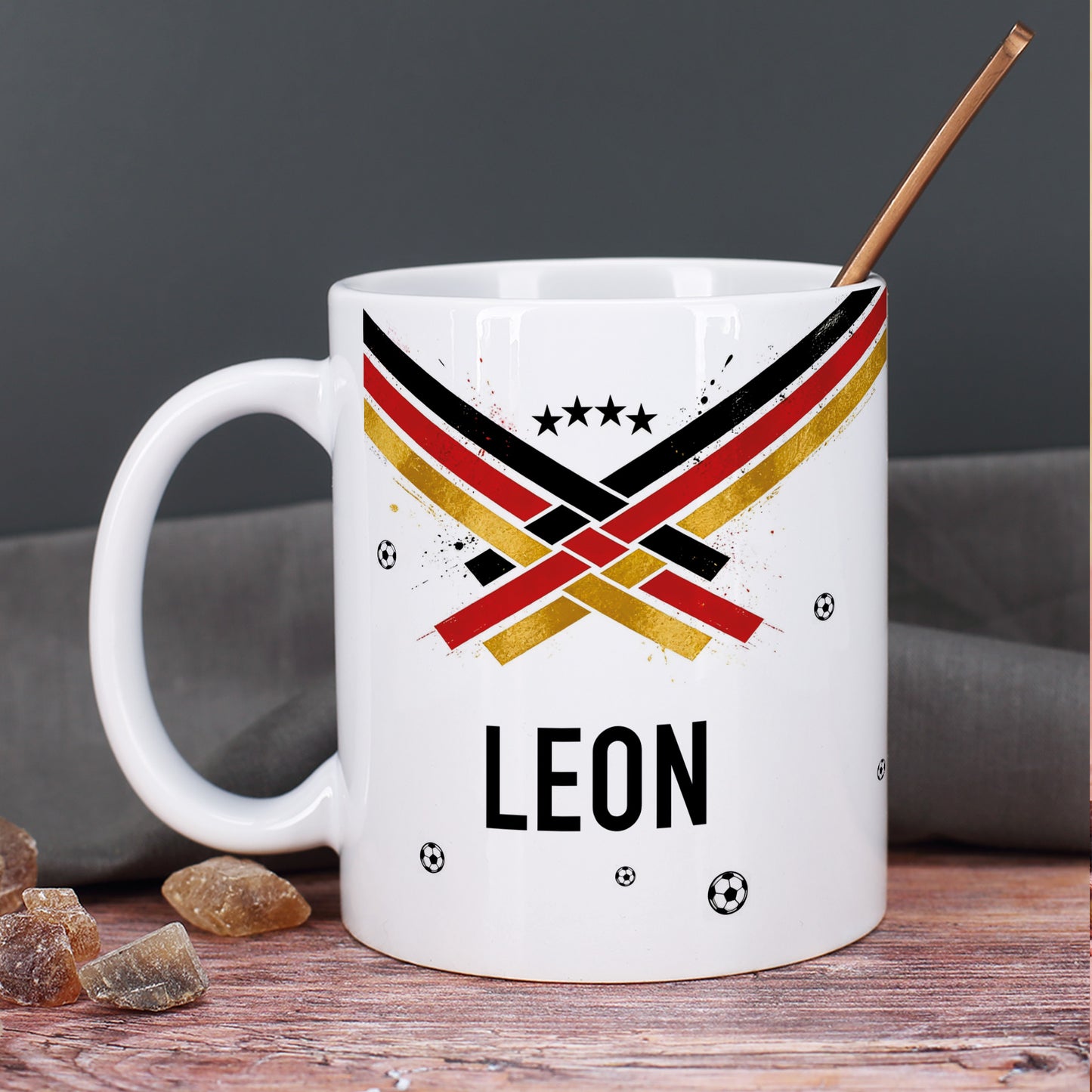 Weiße Keramiktasse mit Fußball Fan-Motiv in Schwarz Rot Gold, kleinen Bällen und Kupferlöffel im Büro