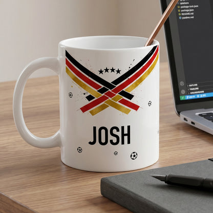 Weiße Fußballtasse mit Name Josh, schwarz-rot-goldenem Motiv, kleinen Bällen und Laptop auf dem Schreibtisch
