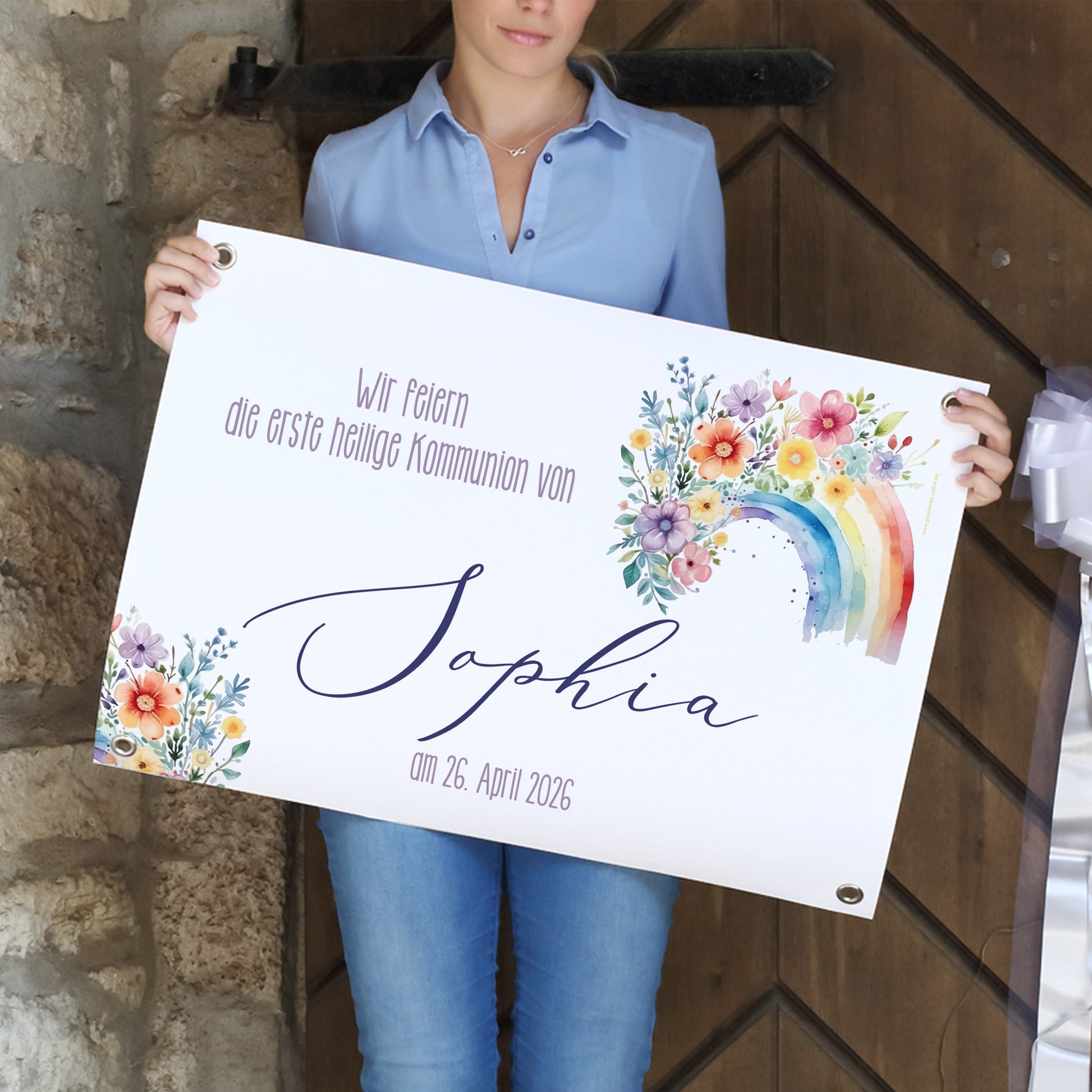 Frau hält ein personalisiertes Banner zur Kommunion in den Händen. Darauf steht: „Wir feiern die erste heilige Kommunion von Sophia – am 26. April 2026“. Das Banner zeigt einen Regenbogen aus Aquarellfarben, umgeben von einem floralen Blumenmotiv in fröhlichen Farben. Die Ecken des Banners sind mit Metallösen versehen.