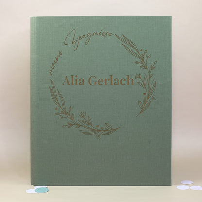 Grüne Zeugnismappe aus Leinen mit floralen Illustrationen im Ring, Aufdruck „meine Zeugnisse“ und personalisiertem Namen „Alia Gerlach“.