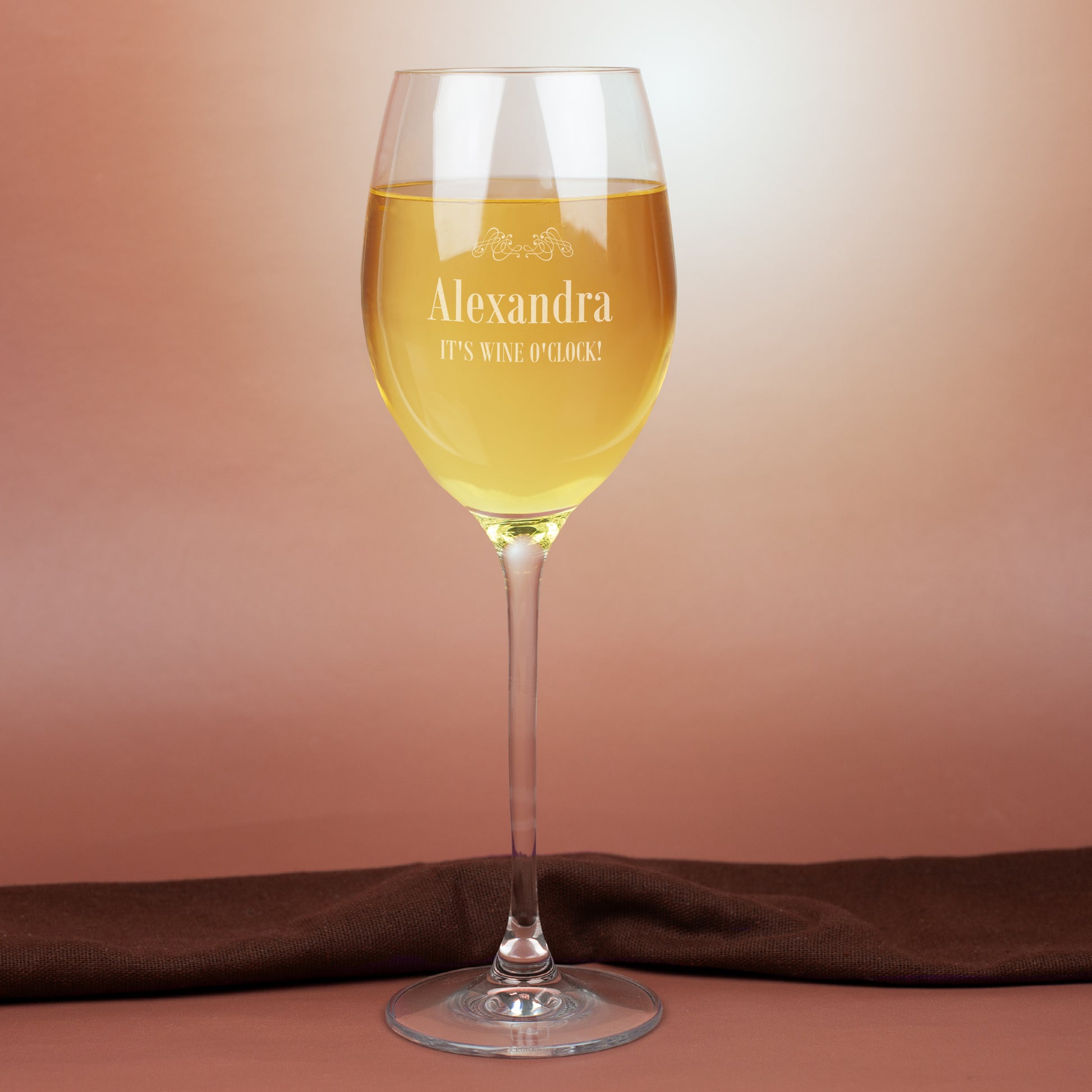 Leonardo Weißweinglas mit Gravur Alexandra und Spruch It’s Wine O’Clock, gefüllt mit Weißwein, auf warmem Hintergrund mit Stoffdeko