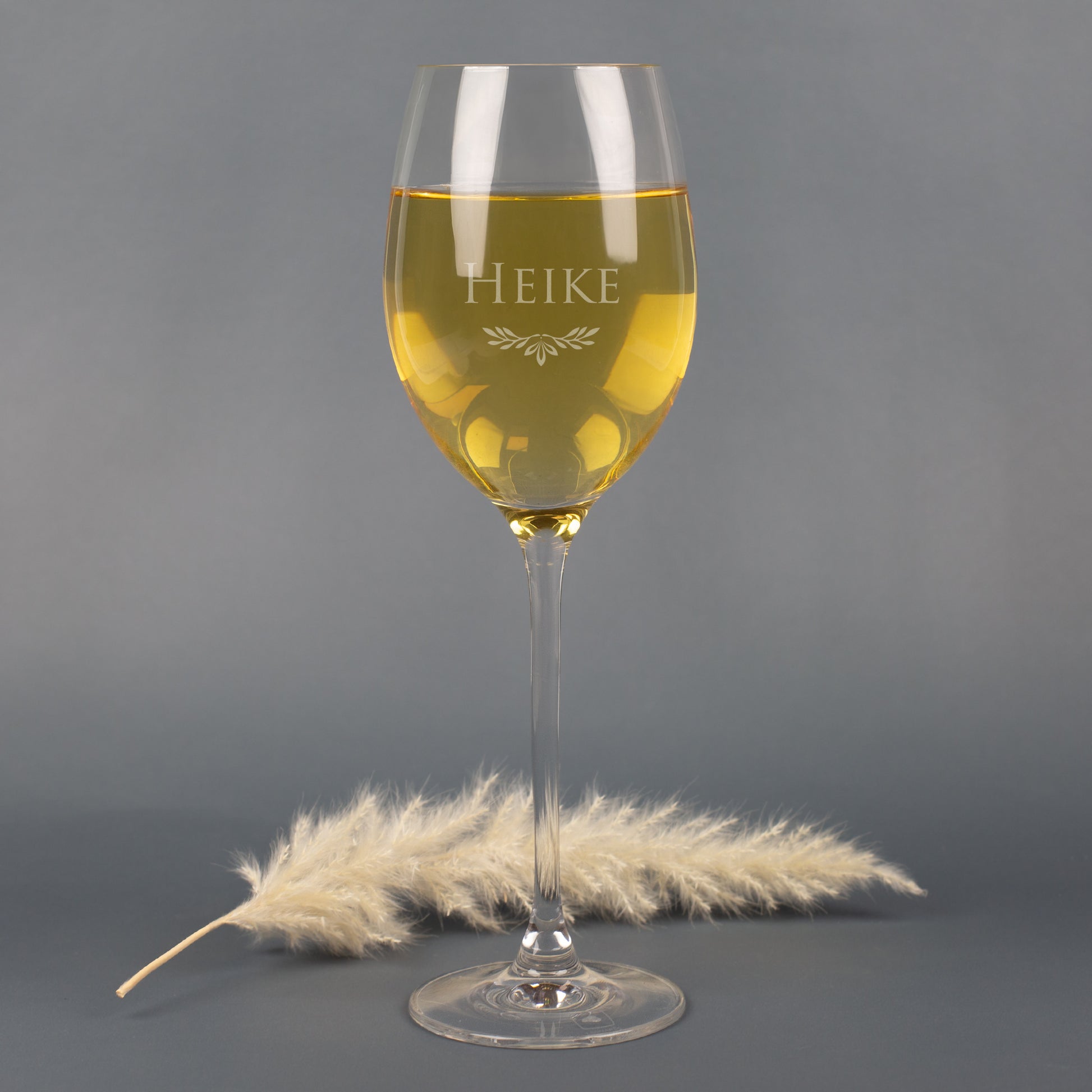 Leonardo Weißweinglas mit Gravur Name Heike und Ornamentmotiv, gefüllt mit Weißwein, auf grauem Hintergrund mit Pampasgras