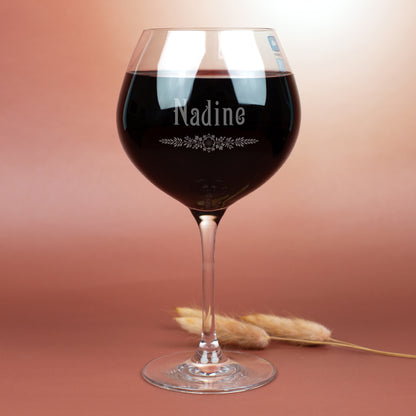 Leonardo Burgunderglas mit Gravur des Namens Nadine und filigranem Ornamentmotiv – elegantes Weinglas mit klarer Gravur, gefüllt mit Rotwein, fotografiert vor braunem Hintergrund mit Trockenblumen.