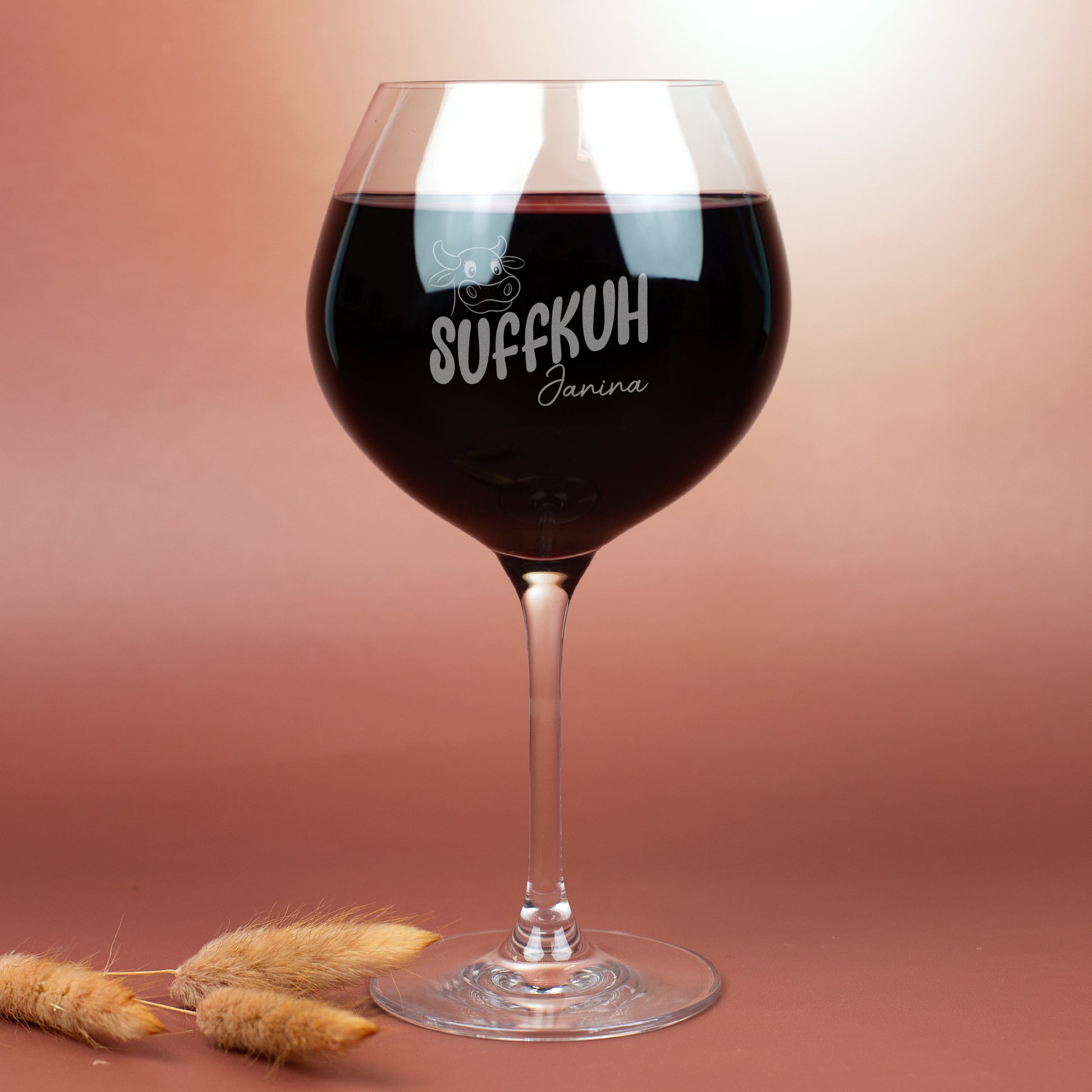 Burgunderglas von Leonardo mit Gravur „Suffkuh“ und Name Janina, gefüllt mit Rotwein – humorvolles Weinglas mit Kuh-Motiv, fotografiert auf braunem Hintergrund mit Trockenblumen.