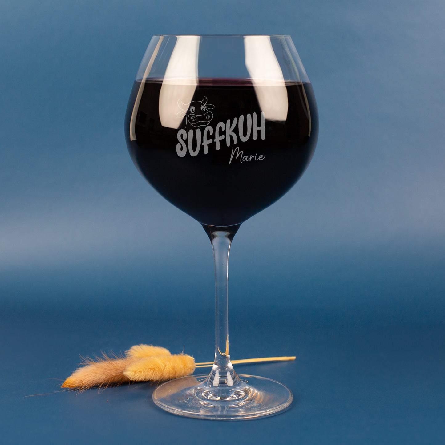 Personalisiertes Leonardo Weinglas mit Gravur „Suffkuh“ und Name Marie – witziges Burgunderglas aus klarem Glas, gefüllt mit Rotwein, vor blauem Hintergrund mit Deko-Halmen.