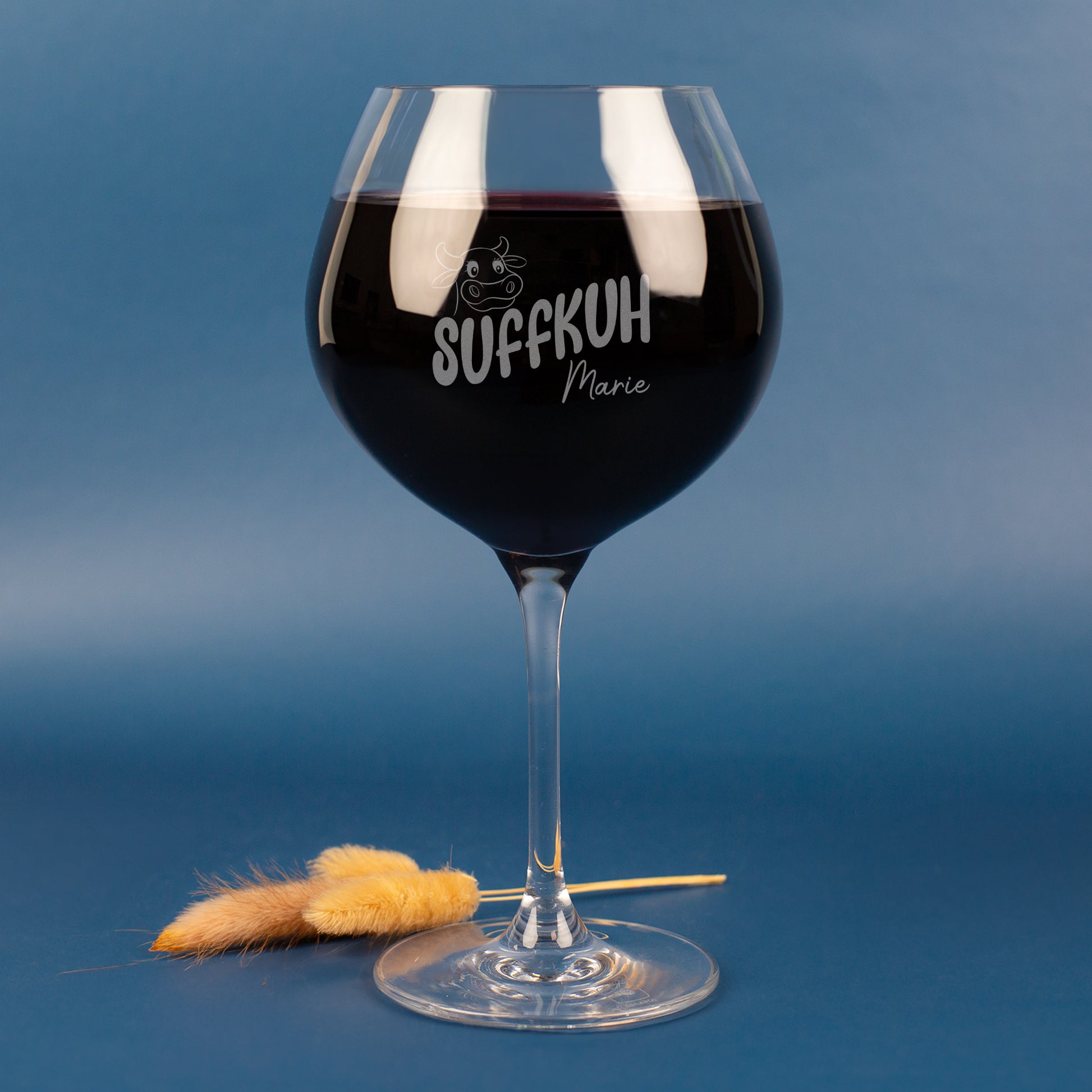 Personalisiertes Leonardo Weinglas mit Gravur „Suffkuh“ und Name Marie – witziges Burgunderglas aus klarem Glas, gefüllt mit Rotwein, vor blauem Hintergrund mit Deko-Halmen.
