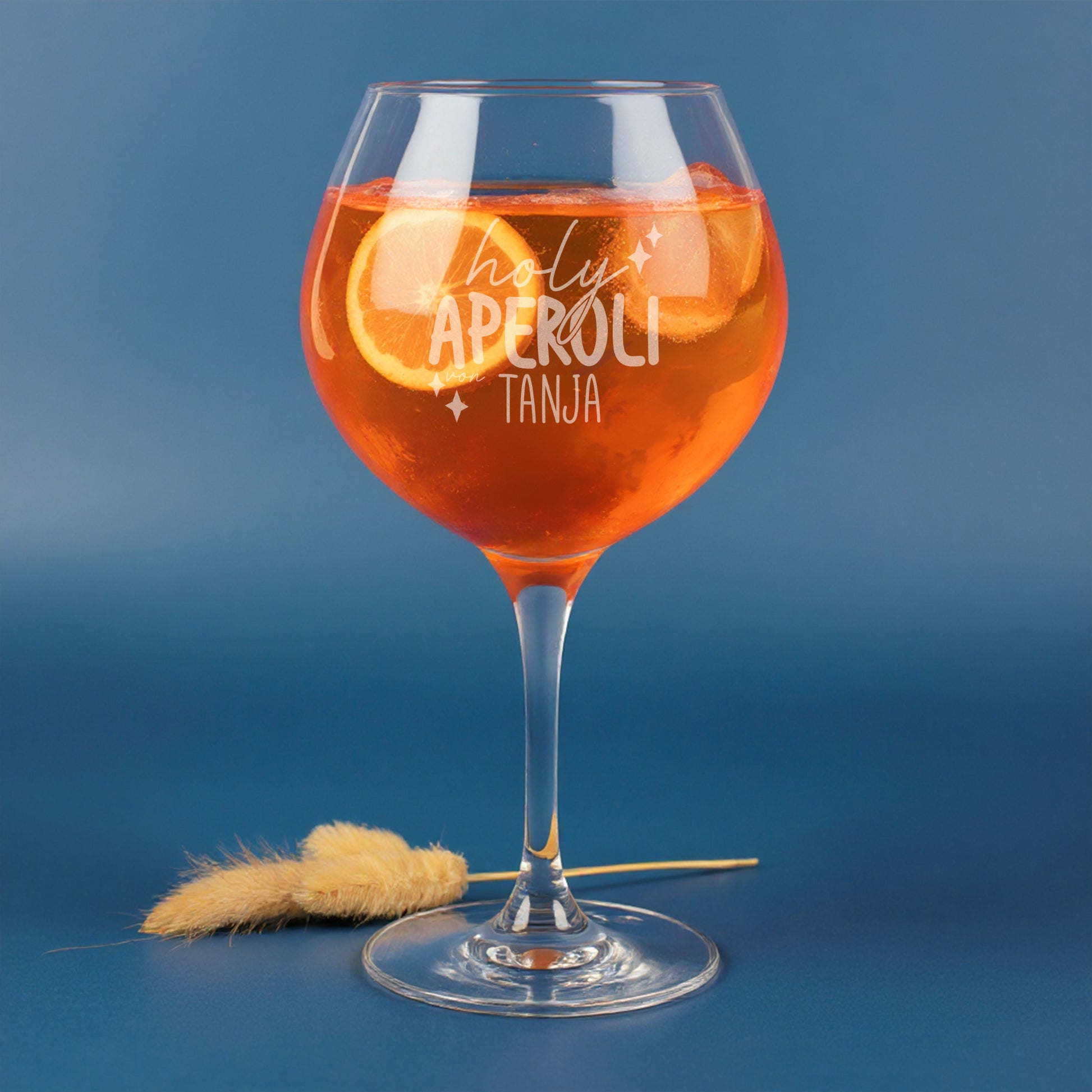 Personalisierte Gravur „holy aperoli von Tanja“ auf Leonardo Weinglas, gefüllt mit Aperitifgetränk, Orangenscheiben und Eiswürfeln – modernes Glas mit witzigem Spruch vor blauem Hintergrund.