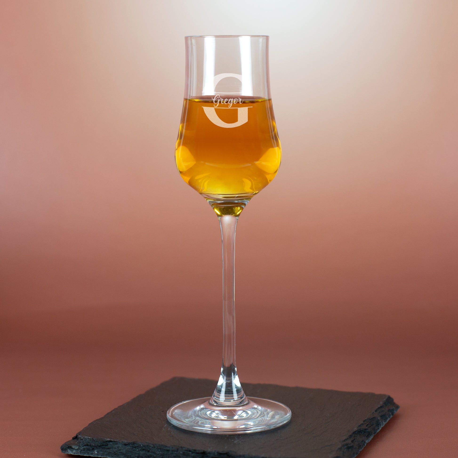 Personalisiertes Grappaglas mit Gravur „Gregor“ und Initiale G – auf Schieferplatte vor rötlichem Hintergrund.
