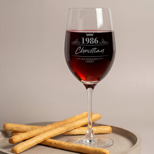 Leonardo Weinglas mit Gravur „1986 Christian – Zur Perfektion gereift“, mit Rotwein gefüllt auf einem Tablett mit Gebäck