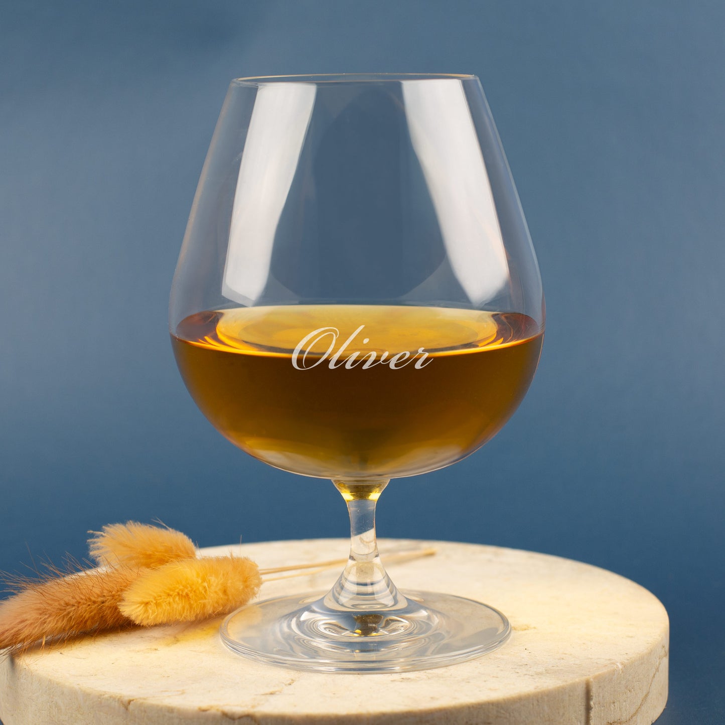 Leonardo Cognacglas mit Gravur „Oliver“ – auf hellem Holzuntergrund mit Trockenblumen, blauer Hintergrund.