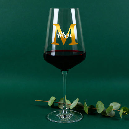 Personalisiertes Leonardo Weinglas mit goldener Initiale M und Name Maxi, gefüllt mit Rotwein vor grünem Hintergrund