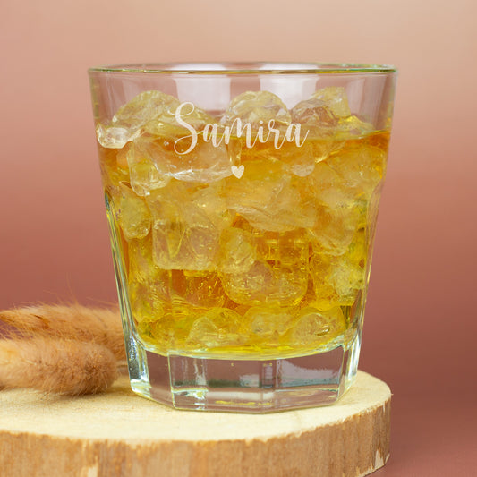 Leonardo-Trinkglas mit Gravur von Name und Herzmotiv, gefüllt mit goldfarbenem Getränk und Eiswürfeln, auf dunkler Schieferplatte vor warmem Hintergrund – elegantes, personalisiertes Glas mit zarter Gravur.