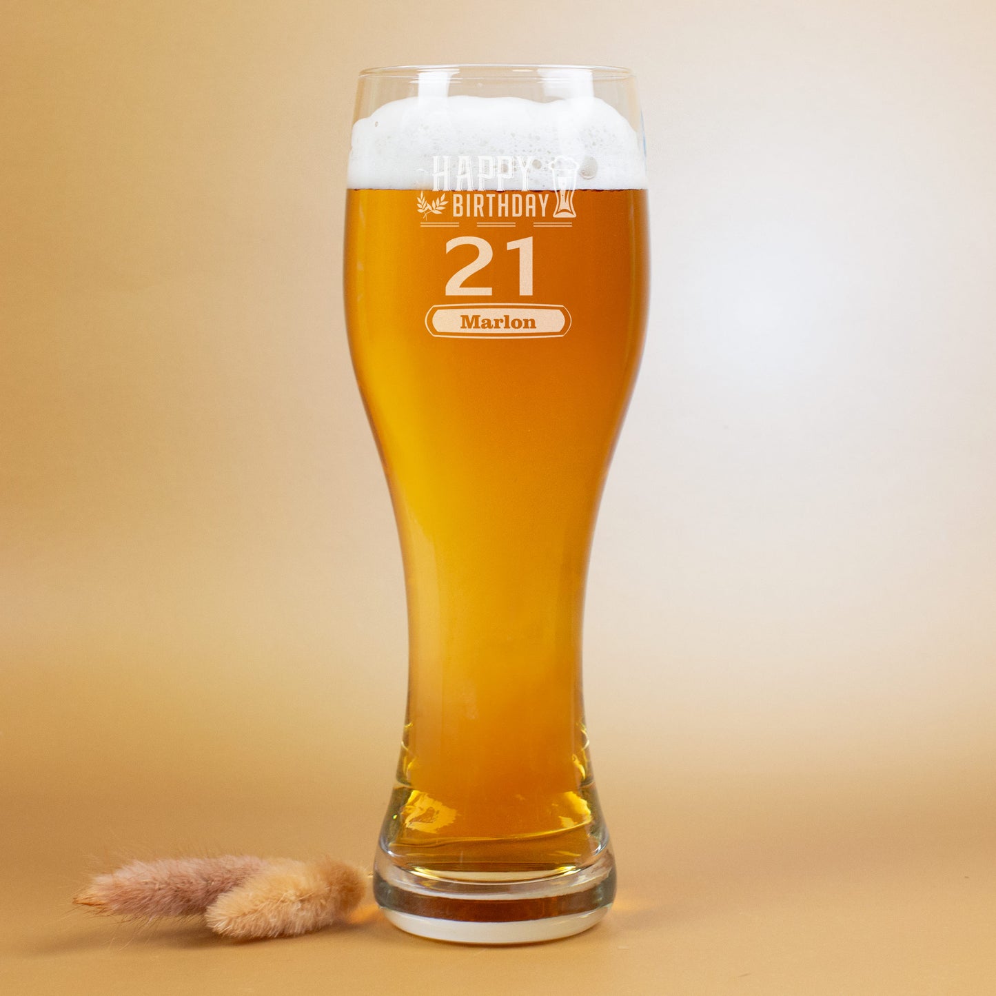 Ein gefülltes Weizenglas mit Schaumkrone, auf dem „Happy Birthday“ sowie die Zahl 21 und der Name „Marlon“ eingraviert sind. Das Glas steht vor beigem Hintergrund, daneben liegt ein kleiner Trockenblumen-Zweig.