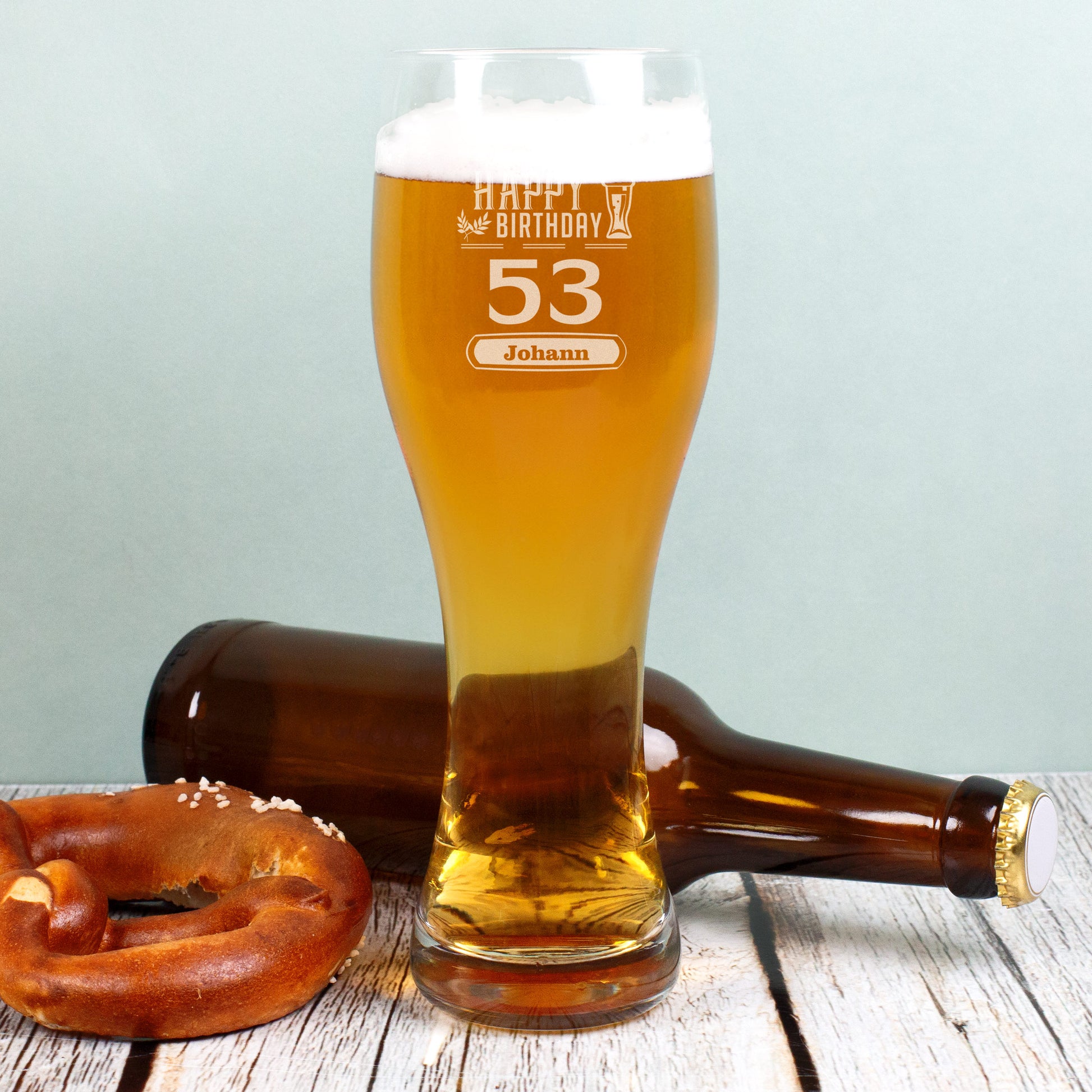 Ein gefülltes Weizenglas mit Schaum, graviert mit „Happy Birthday“, der Zahl 53 und dem Namen „Johann“. Daneben liegen eine Brezel und eine leere Bierflasche, Hintergrund ist ein hellblaues Holzpaneel.