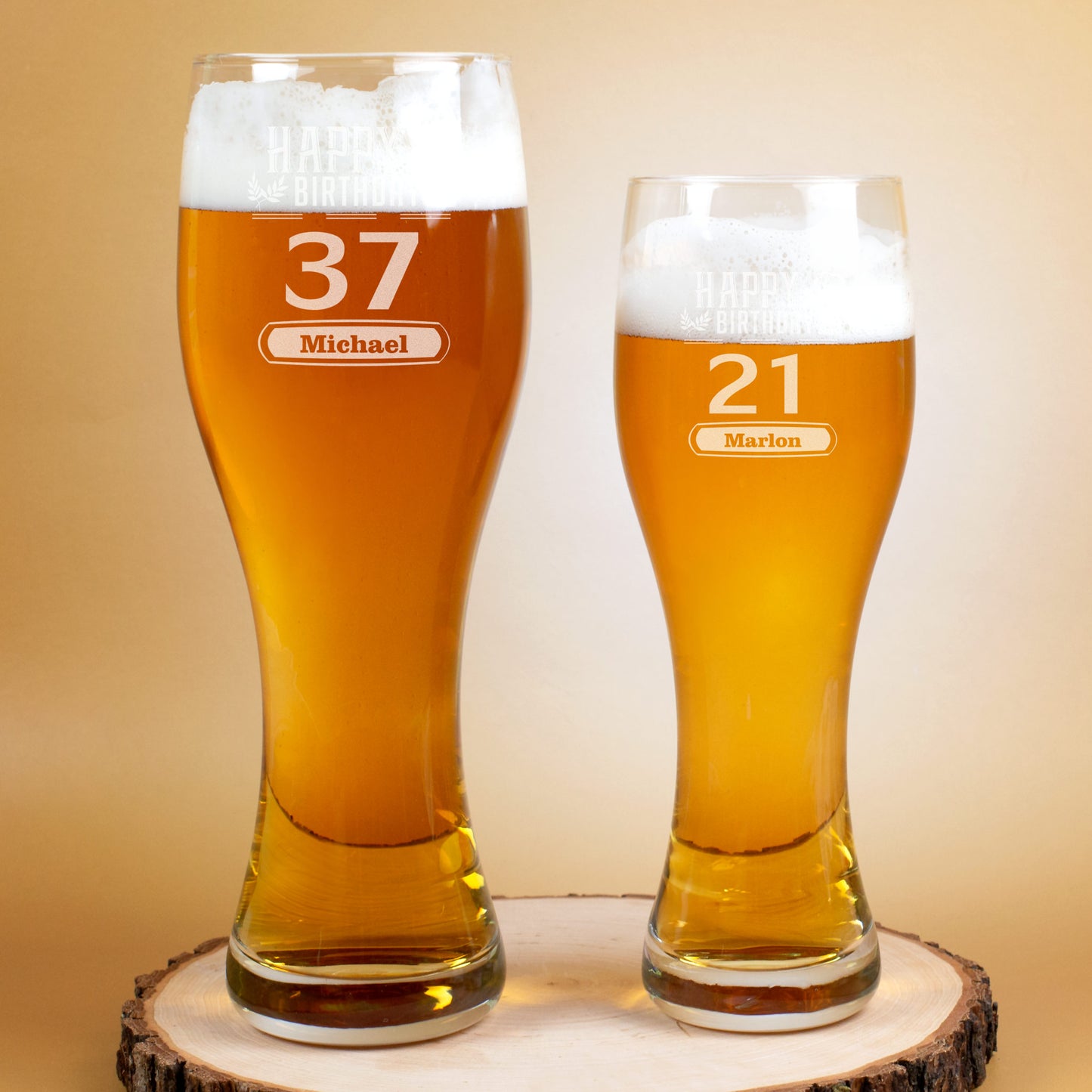 Zwei nebeneinander stehende gefüllte Weizengläser mit Gravur. Das linke zeigt „Happy Birthday“, die Zahl 37 und den Namen „Michael“, das rechte zeigt die Zahl 21 und den Namen „Marlon“. Beide Gläser stehen auf einer runden Holzscheibe.