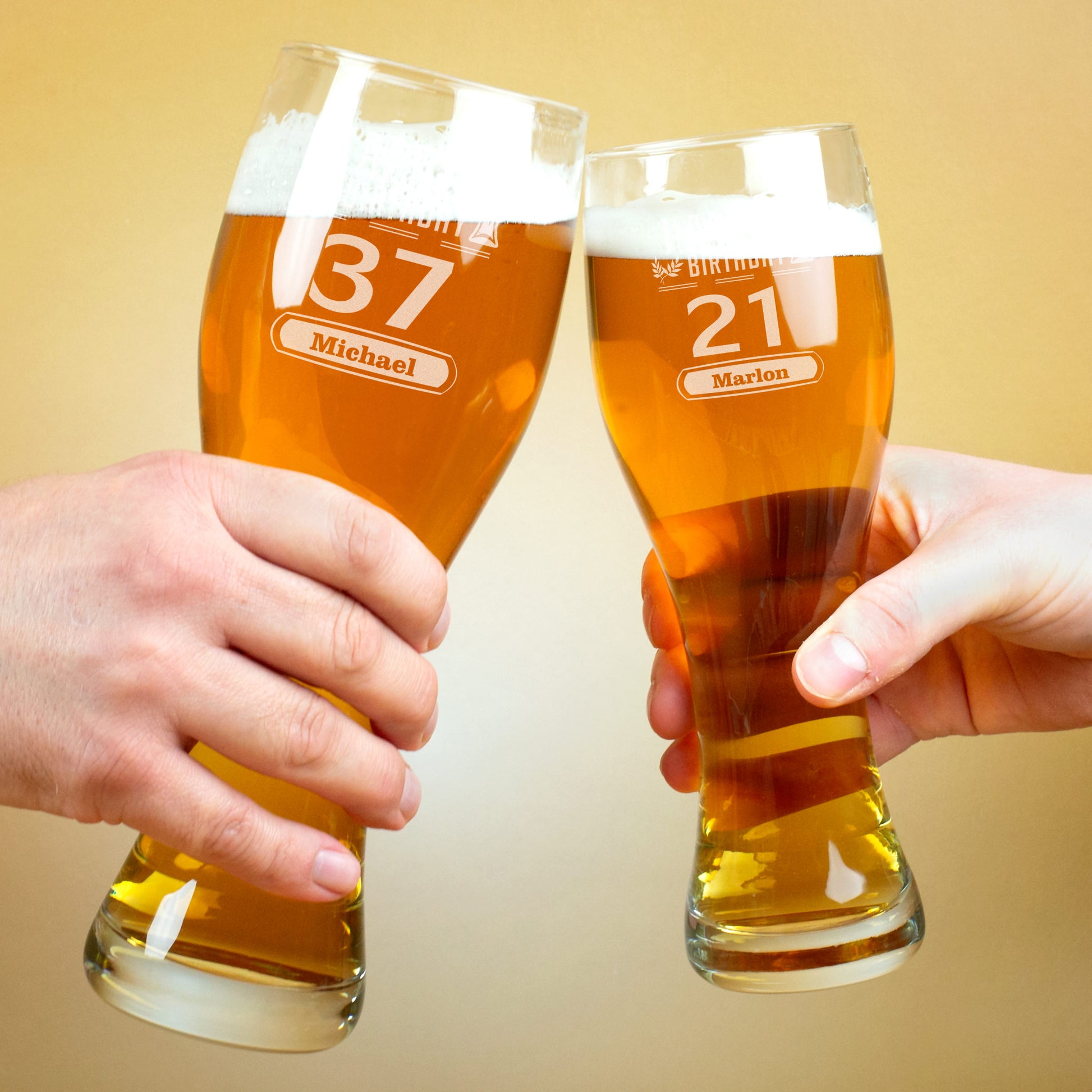 Zwei Hände stoßen mit gefüllten gravierten Weizengläsern an. Links ist das Glas mit der Zahl 37 und dem Namen „Michael“ zu sehen, rechts das Glas mit der Zahl 21 und dem Namen „Marlon“.