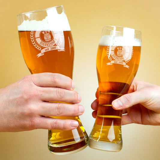 Zwei gravierte Biergläser im Anstoßen, beide gefüllt mit hellem Bier, Gravur „Bierkenner“ erkennbar