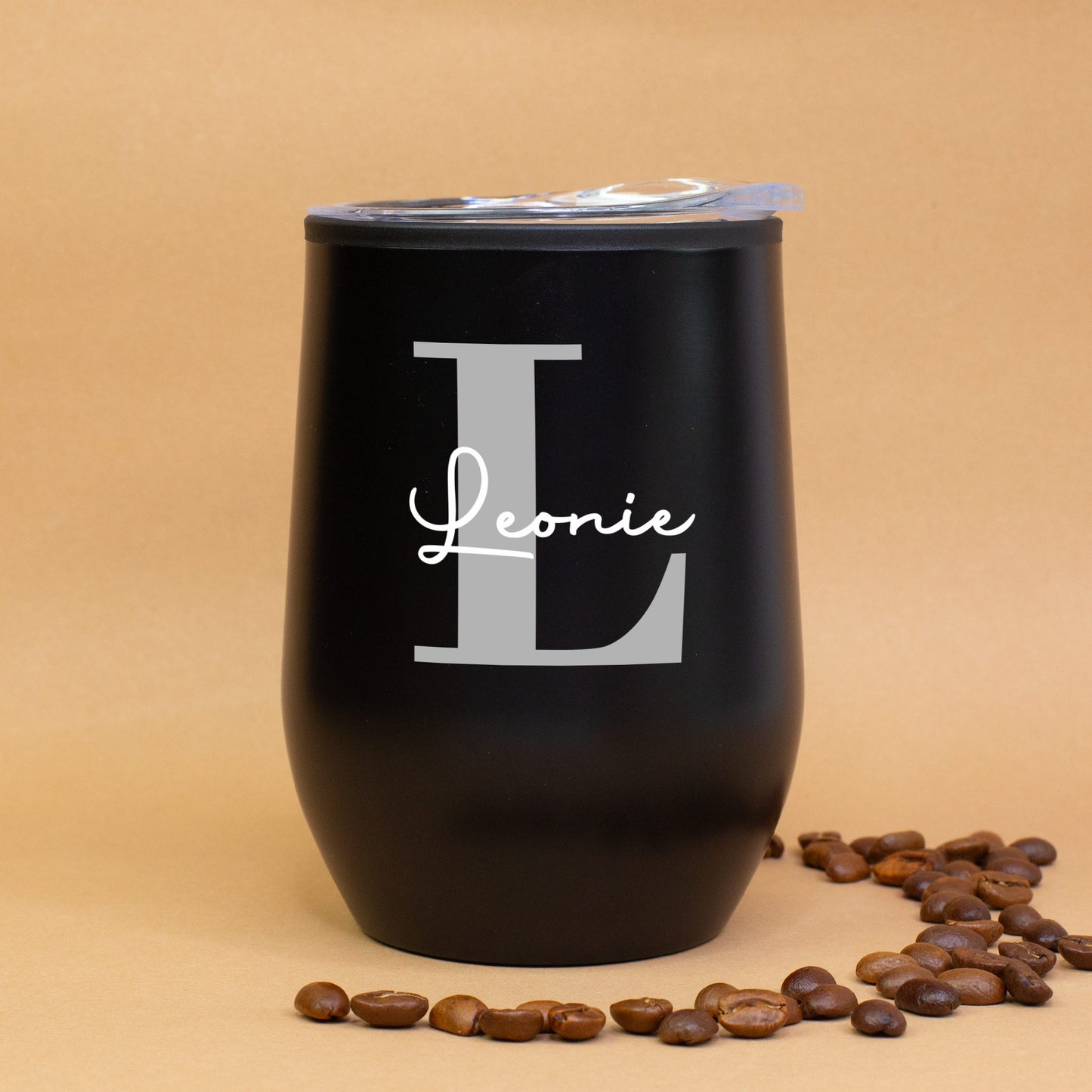 Schwarzer Thermobecher mit Initiale L und Name Leonie, auf braunem Hintergrund mit Kaffeebohnen dekoriert