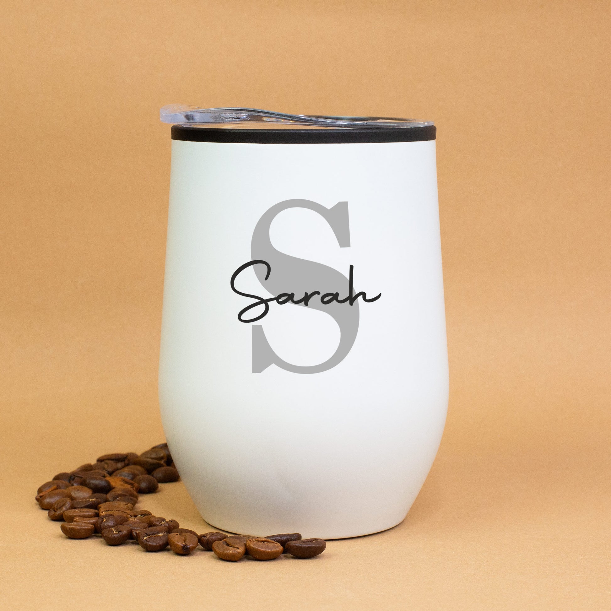 Weißer Thermobecher mit Initiale S und Name Sarah, auf braunem Hintergrund mit Kaffeebohnen dekoriert