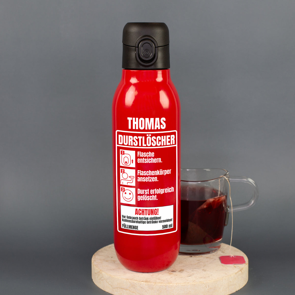 Personalisierte rote Trinkflasche mit Aufdruck „Thomas Durstlöscher“ neben einem Glas Tee