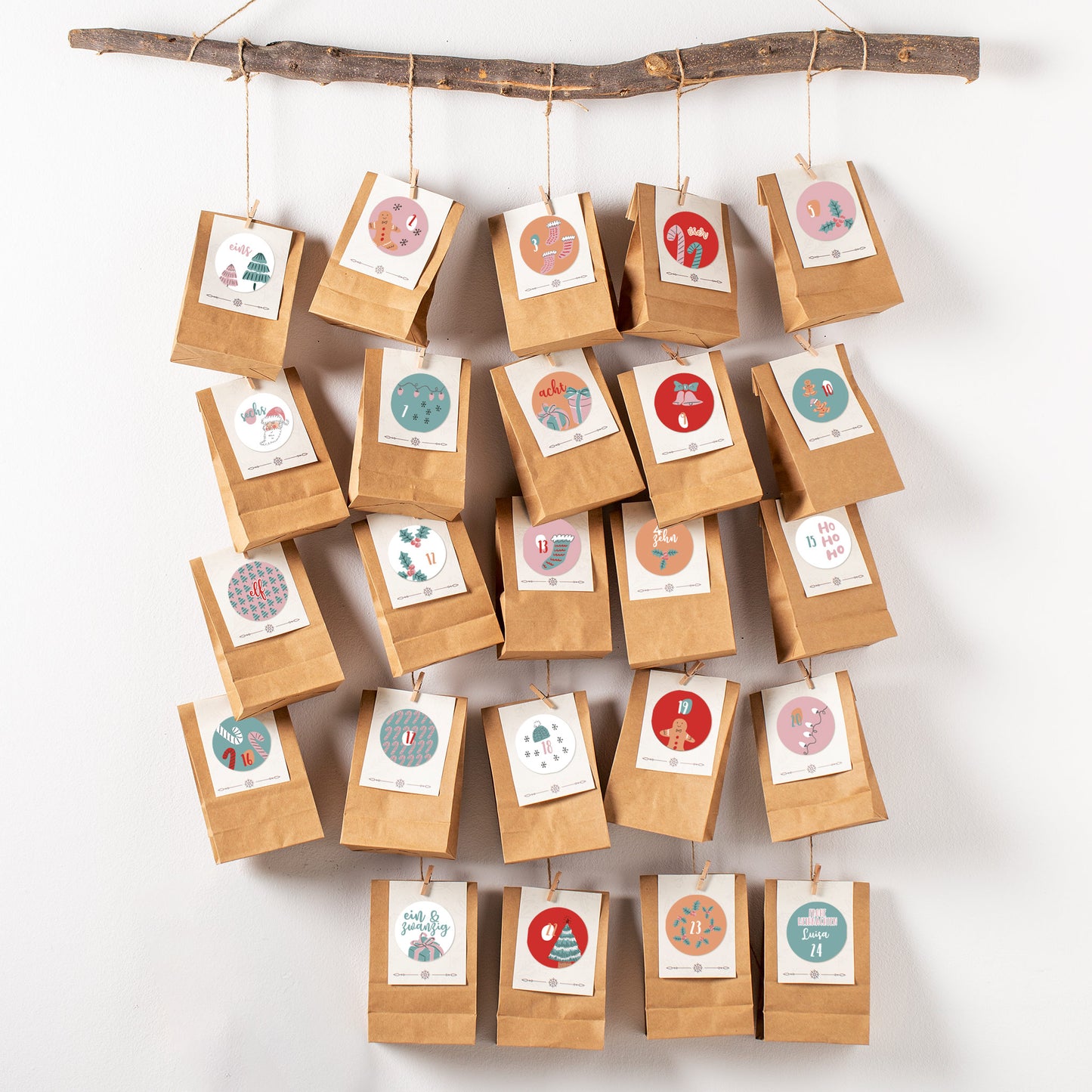DIY-Adventskalender aus Papiertüten mit personalisierten rosa-türkisen Aufklebern, aufgehängt an einem Ast