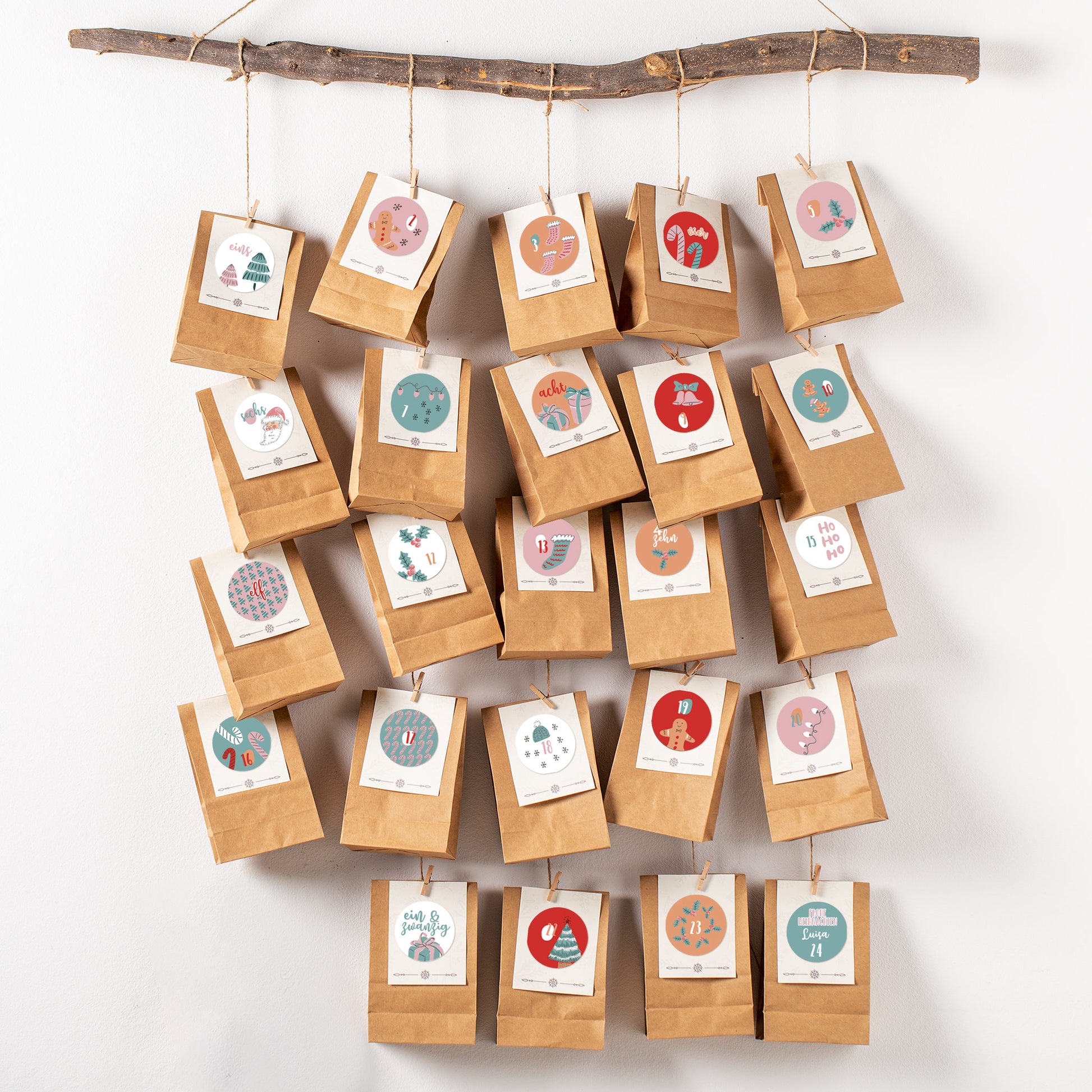 DIY-Adventskalender aus Papiertüten mit personalisierten rosa-türkisen Aufklebern, aufgehängt an einem Ast