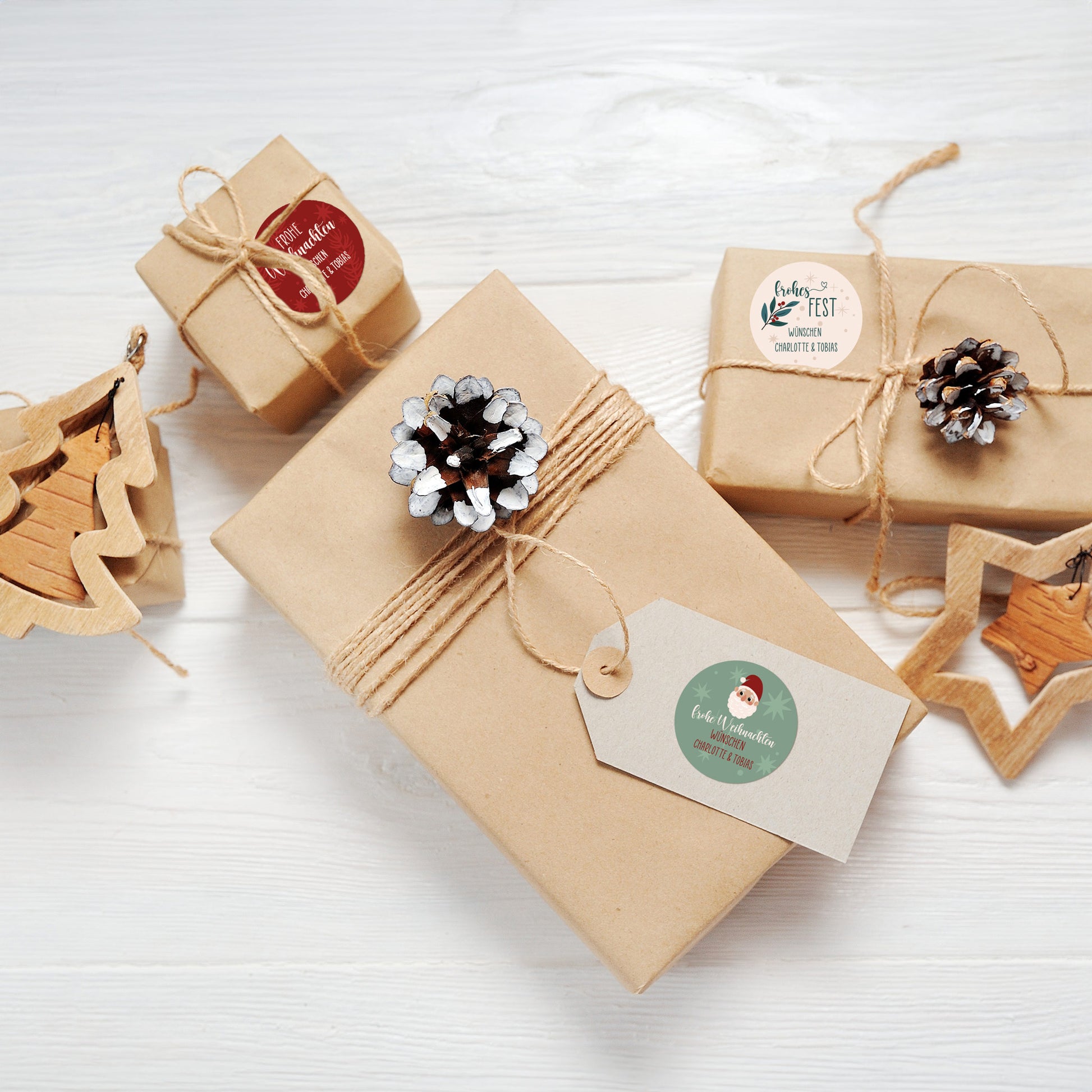 Geschenke im Naturlook mit Juteschnur und Zapfen, dekoriert mit Aufklebern „Frohes Fest“ und „Frohe Weihnachten“ sowie Santa-Motiv auf Etikett.