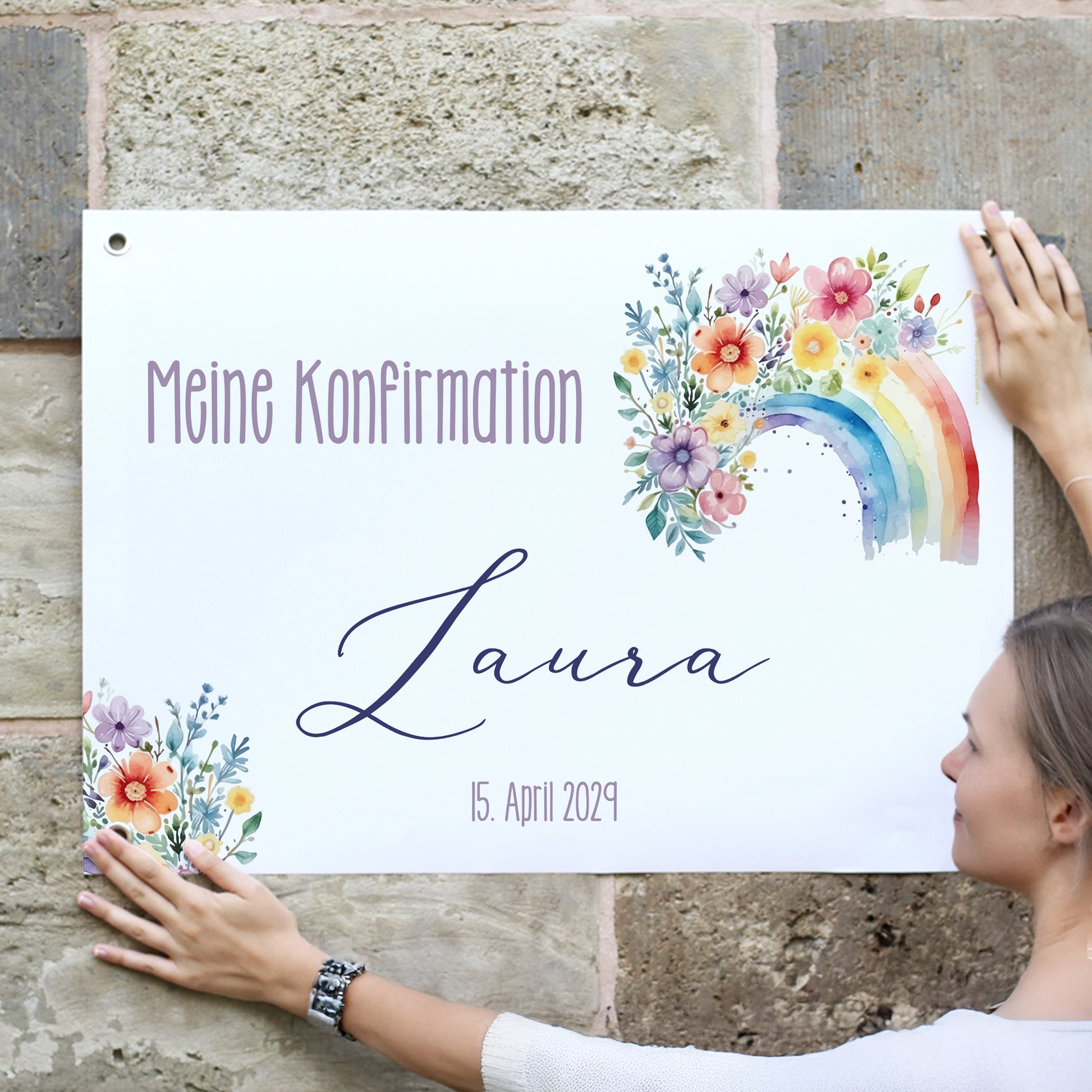 Junge Frau hängt ein personalisiertes Banner an eine Natursteinwand. Text: „Meine Konfirmation – Laura – 15. April 2029“. Das Banner ist mit einem bunten Aquarell-Regenbogen und floralen Blumenillustrationen geschmückt. Die Ecken sind mit stabilen Metallösen versehen.