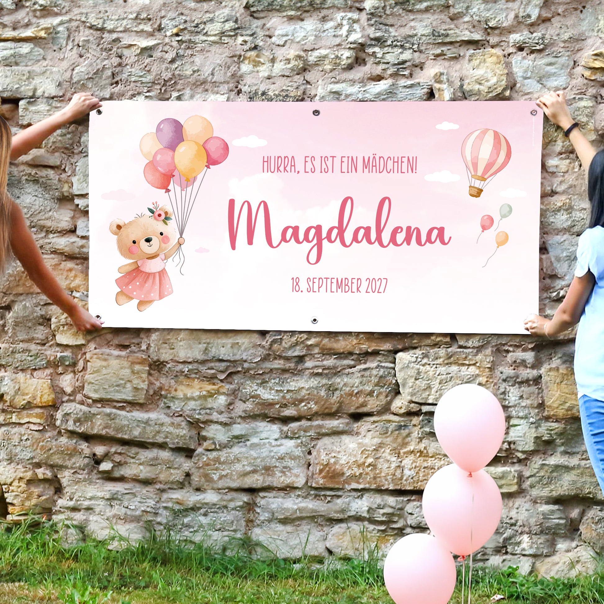 Rosa Geburtsbanner mit Teddy Motiv, Name Magdalena und Wunschtext „Hurra, es ist ein Mädchen“, von zwei Personen an Steinwand gehalten