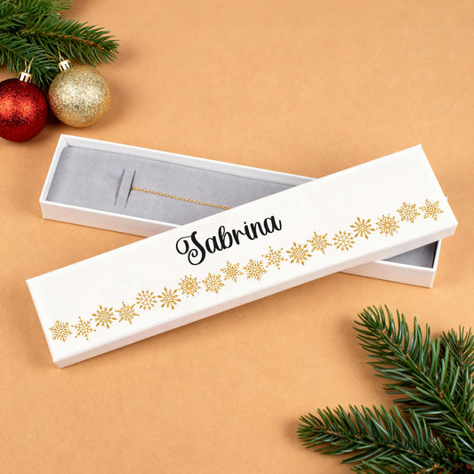 Personalisierte Schmuckverpackung aus weißem Karton mit goldenen Schneeflocken und dem Namen „Sabrina“ in schwarzer Schrift. Der Deckel liegt geöffnet neben der Box und zeigt ein Armband auf grauem Schaumstoffeinsatz. Dekoriert mit Tannenzweigen und roten sowie goldenen Weihnachtskugeln.