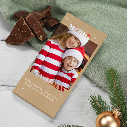 Personalisierte Schokoladentafel zu Weihnachten mit Foto und Wunschtext im „Merry Christmas“-Design. Die Verpackung besteht aus naturfarbenem Kraftpapier und ist mit weißen Sternen verziert. Auf dem Foto sind zwei Kinder mit Weihnachtsmützen abgebildet, daneben liegen Stücke feinster Vollmilchschokolade. Perfektes Geschenk zu Weihnachten, Nikolaus oder zum Wichteln.