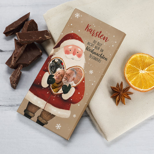 Personalisierte Weihnachts-Schokolade mit Weihnachtsmann und Herzmotiv. Auf der Verpackung ist der Name „Karsten“ aufgedruckt, darunter der Spruch „Du bist nicht nur an Weihnachten besonders“. Im Herzfenster befindet sich ein persönliches Foto. Neben der Schokolade liegen Schokoladenstücke, getrocknete Orangenscheiben und Sternanis – festlich dekoriert auf hellem Hintergrund.