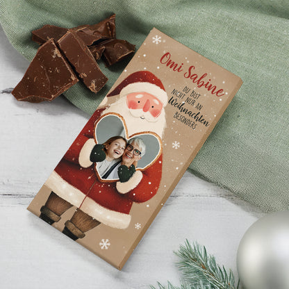Foto-Schokoladentafel mit personalisierter Verpackung im Weihnachtsdesign. Der Weihnachtsmann hält ein Herz mit integriertem Foto, darüber steht „Omi Sabine“. Das Motiv ist von Schneeflocken umgeben, auf dem Tisch liegen Schokoladenstücke, Tannenzweige und eine silberne Kugel. Ideal als Geschenk zu Weihnachten oder Nikolaus.