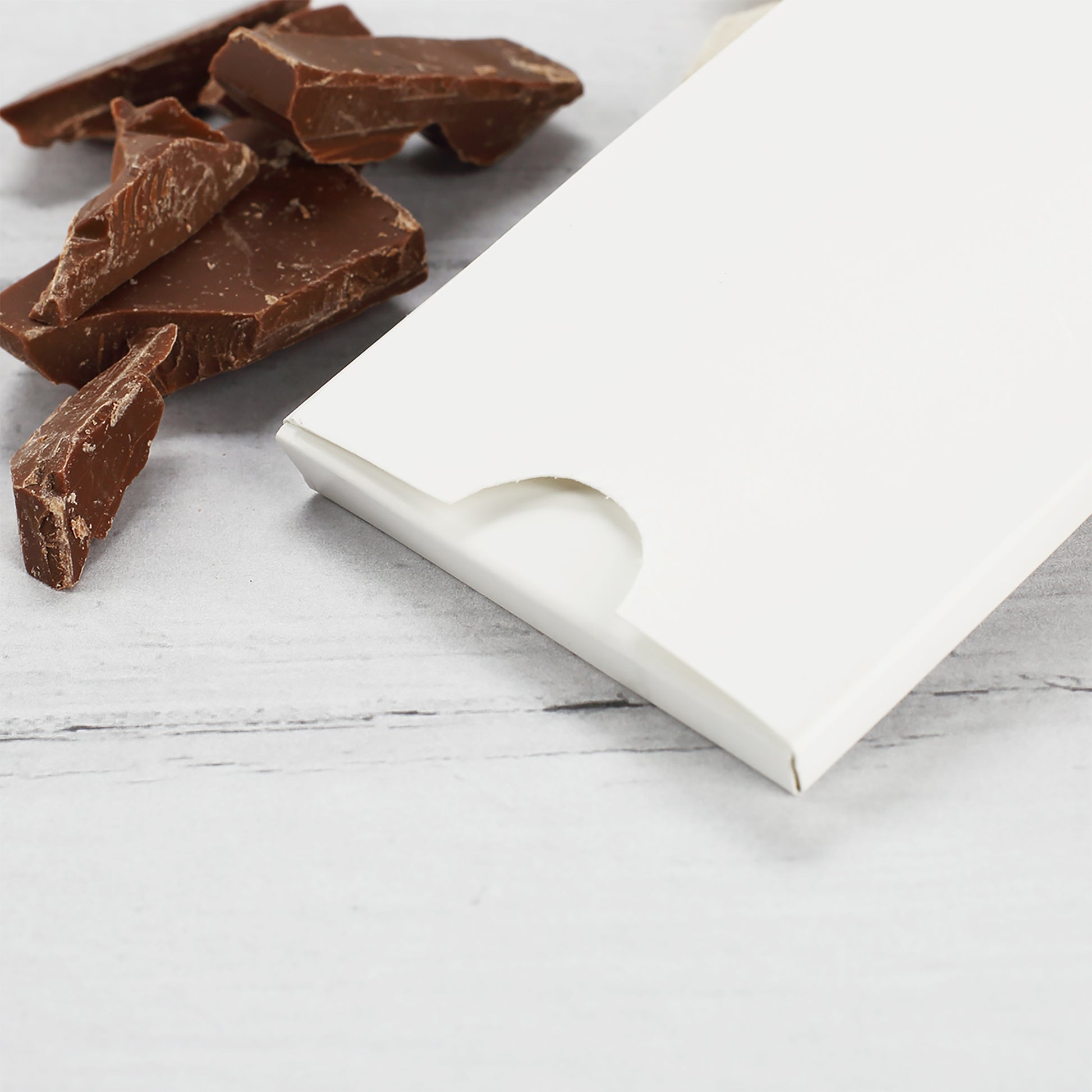 Schokoladenverpackung in Weiß mit Einkerbung zum Öffnen, daneben Schokoladenstücke