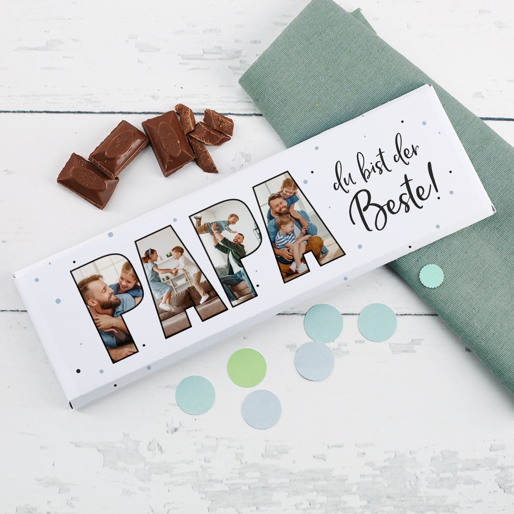 Personalisierbare Fotogeschenk-Schokolade für Papa mit 4 Bildern im Schriftzug „PAPA“ und aufgebrochener Schokolade auf weißem Tisch