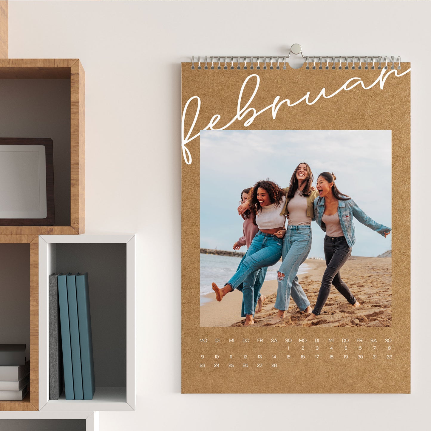 Foto-Wandkalender in A3 mit großformatigem Februar-Foto von vier Freundinnen am Strand, Kraftpapier-Design und Monatsübersicht unten.