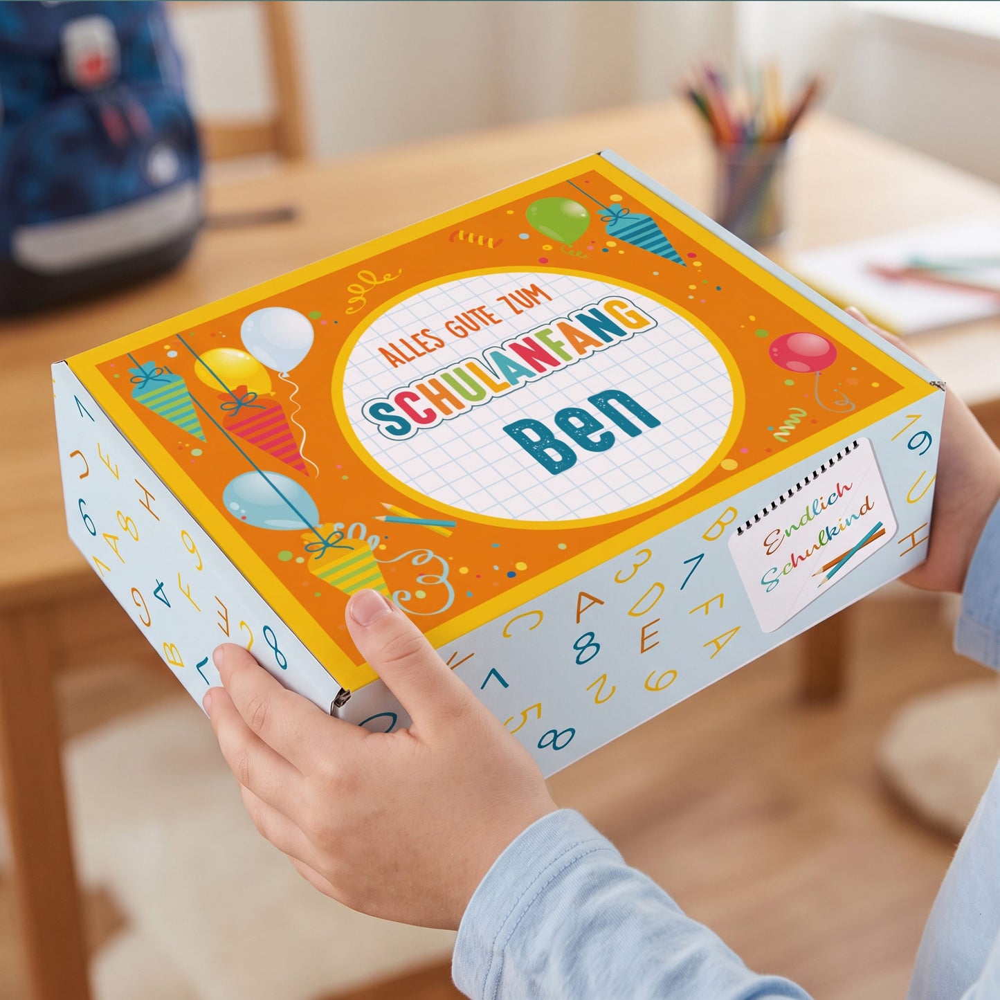 Kleine personalisierte Aufbewahrungsbox aus Pappe mit Name und buntem Schulanfang-Motiv in Kinderhänden