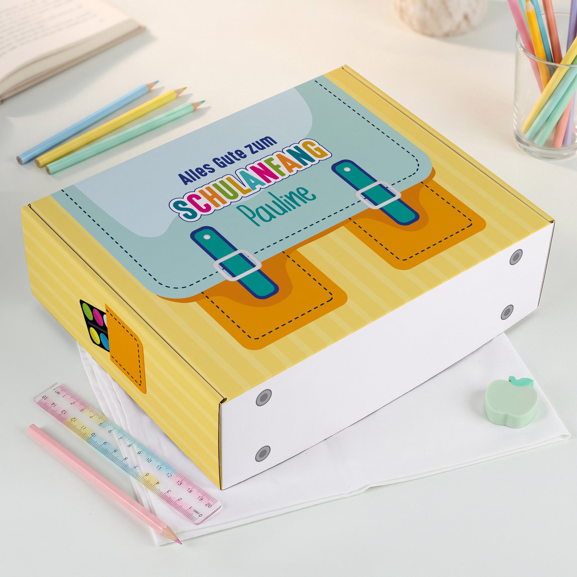 Große gelbe Geschenkbox zur Einschulung mit Name bedruckt, liegend auf einen Schreibtisch