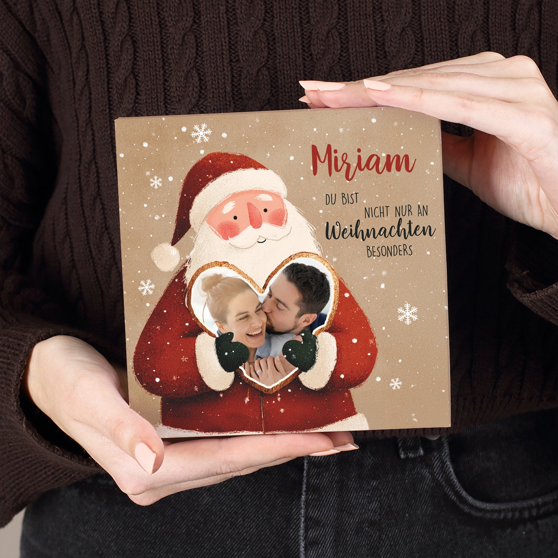 Weihnachtsmann mit freundlichem Gesichtsausdruck, hält Herz mit Foto eines Paares. Darüber personalisierte Beschriftung in roter und schwarzer Schrift. Deutlich sichtbare Struktur des Kartonmaterials und winterliches Schneeflocken-Design.