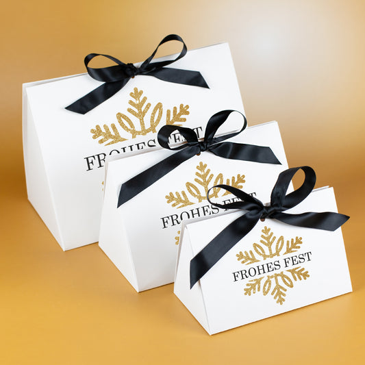 Drei weiße Geschenktaschen in den Größen klein, mittel und groß mit goldfarbener Schneeflocke und schwarzer Satinschleife, im Set vor goldenem Hintergrund präsentiert.