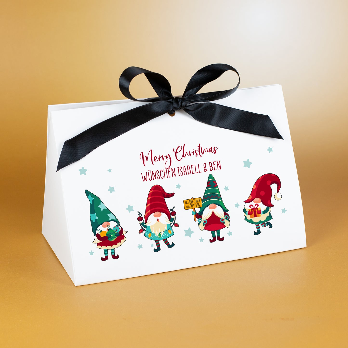 Große Geschenkverpackung zu Weihnachten mit schwarzem Satinband und farbigem Wichtelmotiv. Über dem Motiv steht der personalisierte Text „Merry Christmas wünschen Isabell & Ben“ in roter Schrift. Die vier Wichtel tragen weihnachtliche Outfits und stehen vor einem Muster kleiner Sterne auf weißem Karton. Präsentiert vor goldenem Hintergrund.