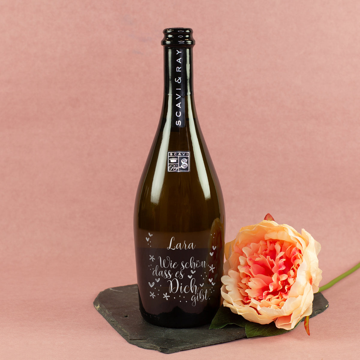 Personalisierte Scavi & Ray Prosecco-Flasche mit Gravur Lara und Spruch Wie schön, dass es Dich gibt neben orangefarbener Blume