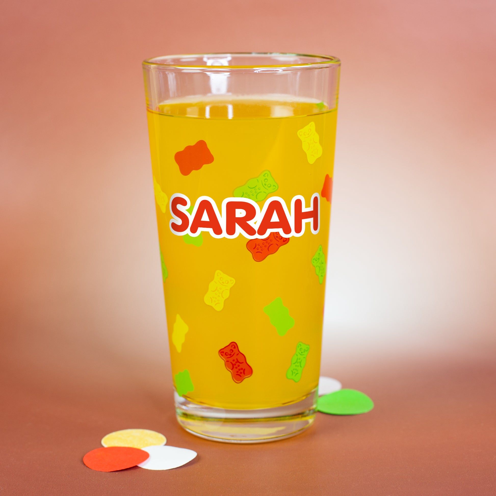 Personalisierbares HARIBO Glas mit bunten Gummibärchenmotiven und großem weißen Namensdruck „Sarah“, gefüllt mit gelber Flüssigkeit.
