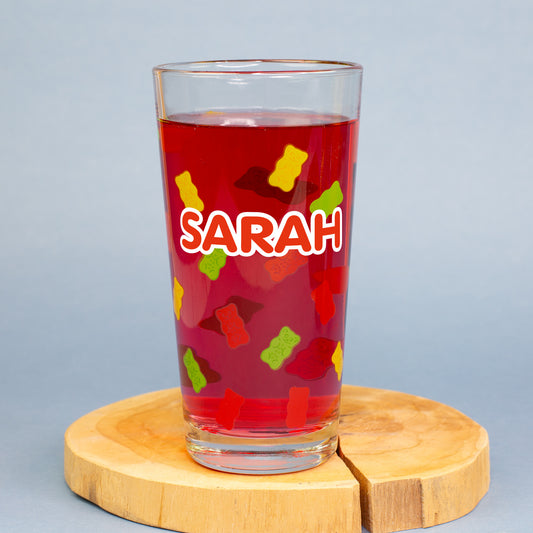 HARIBO Trinkglas mit personalisiertem Aufdruck „Sarah“, gefüllt mit rotem Getränk, bunte Gummibärchenmotive rundum, auf Holzunterlage.