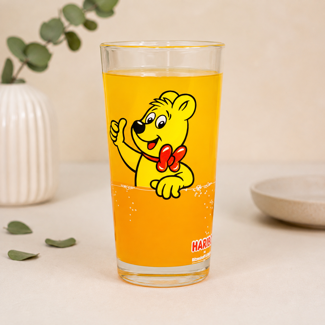 HARIBO Trinkglas ohne Personalisierung mit Goldbär Motiv, orangem Getränk und dezenter Deko auf hellem Hintergrund