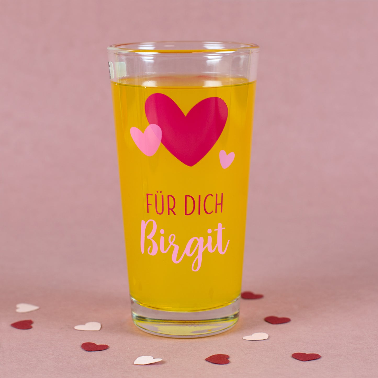 Personalisierbares Glas mit Herz-Motiv und Namen „Birgit“ auf rosa Hintergrund mit Dekoration