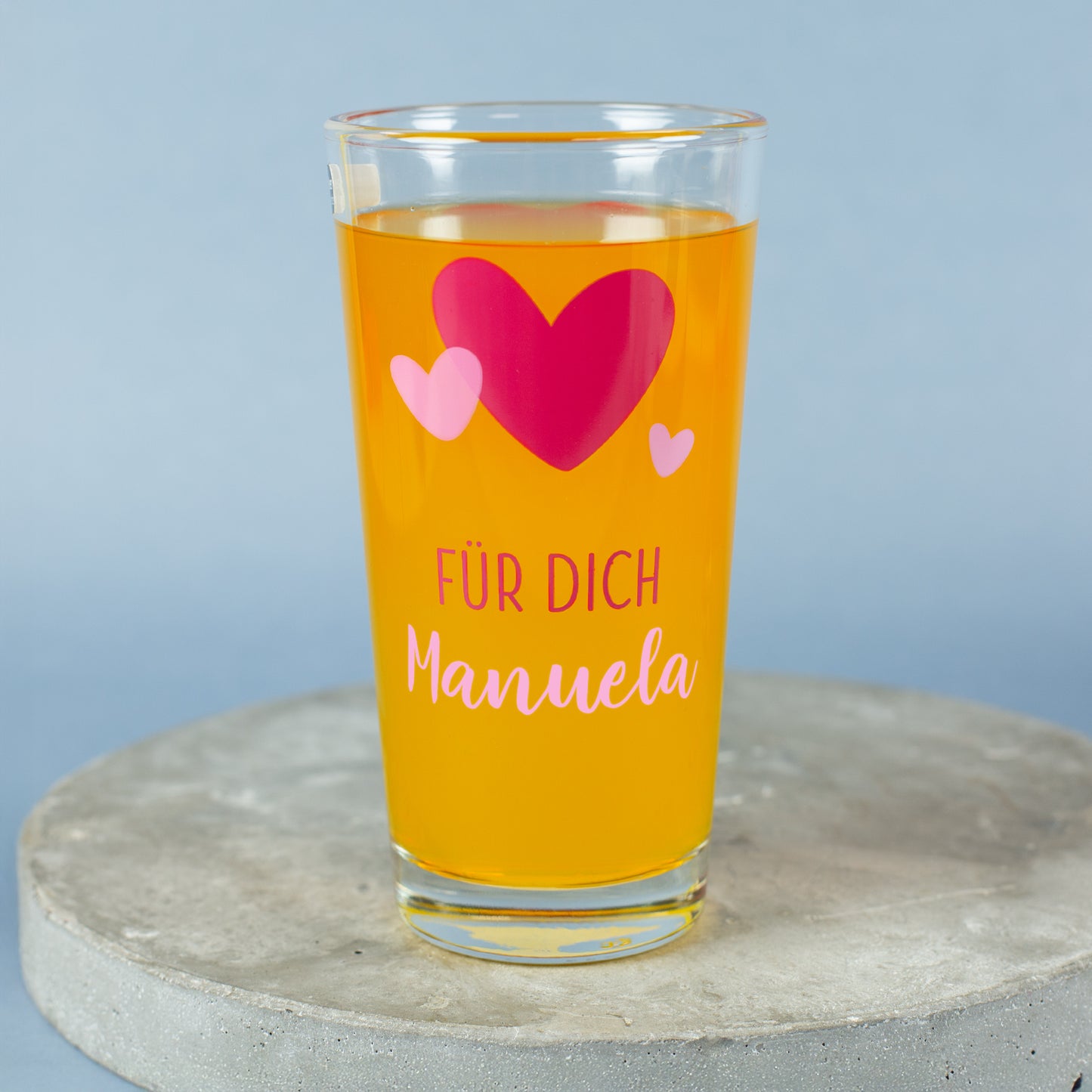 Trinkglas mit Herz-Design und personalisiertem Namen „Manuela“ auf grauem Podest