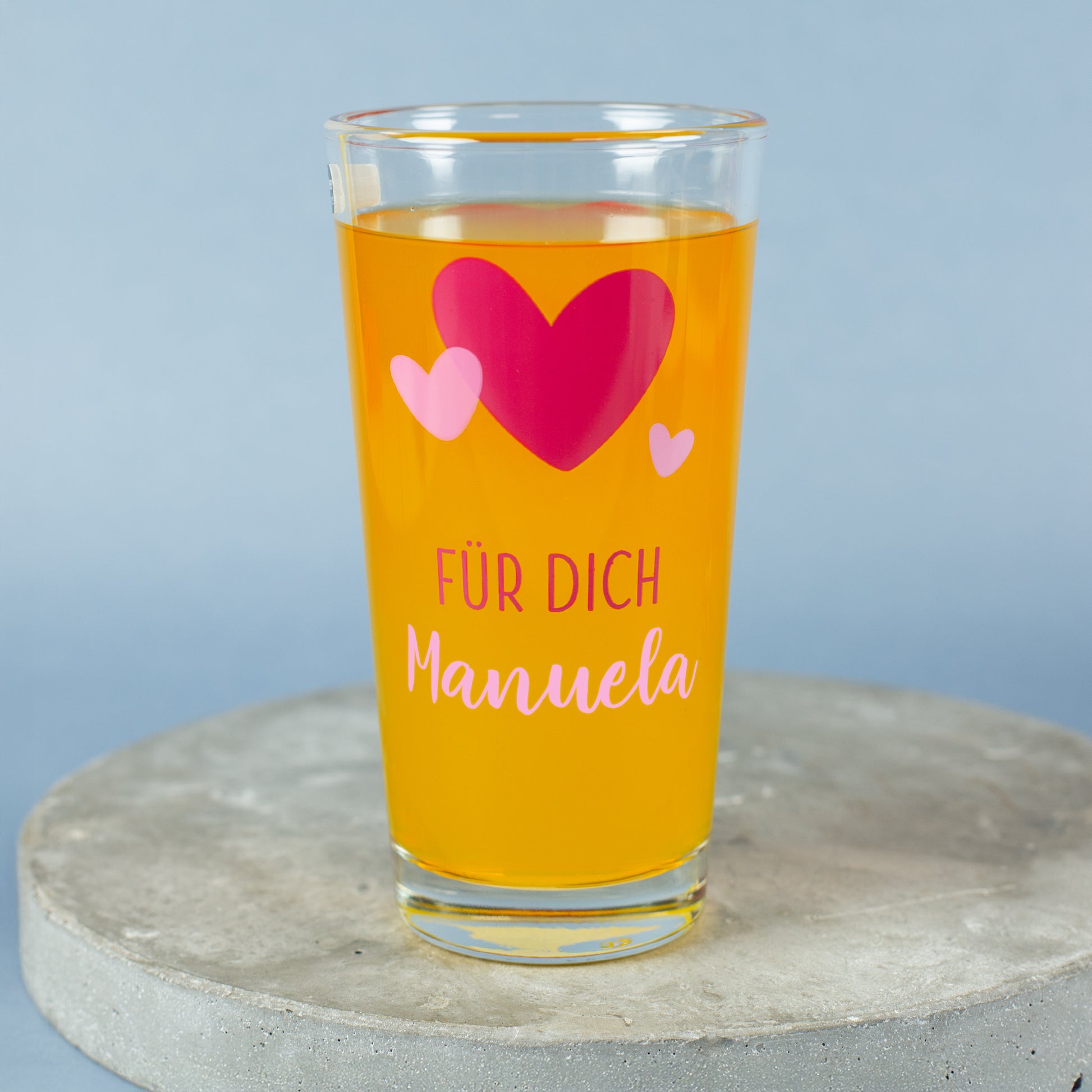 Trinkglas mit Herz-Design und personalisiertem Namen „Manuela“ auf grauem Podest