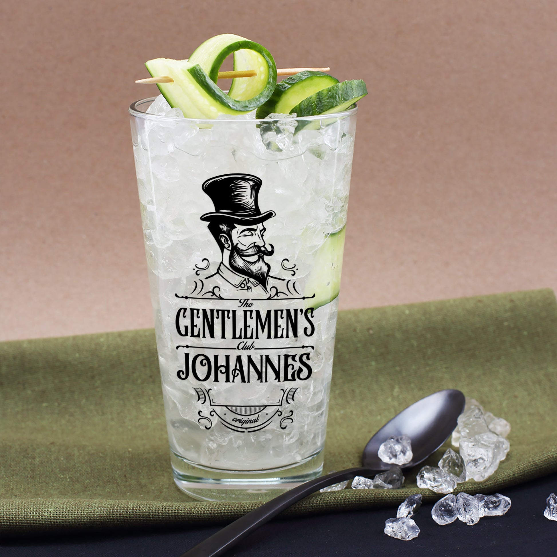 Personalisierbares Trinkglas mit schwarzem Aufdruck „The Gentlemen’s Club“ und Name Johannes, gefüllt mit Eis und Gurkenspiralen